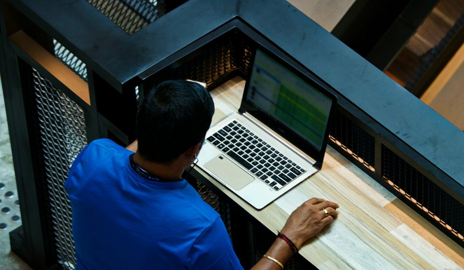 A man with a blue t shirt on and dark hair sitting at a desk with his laptop open, with a blurry spreadsheet on screen.