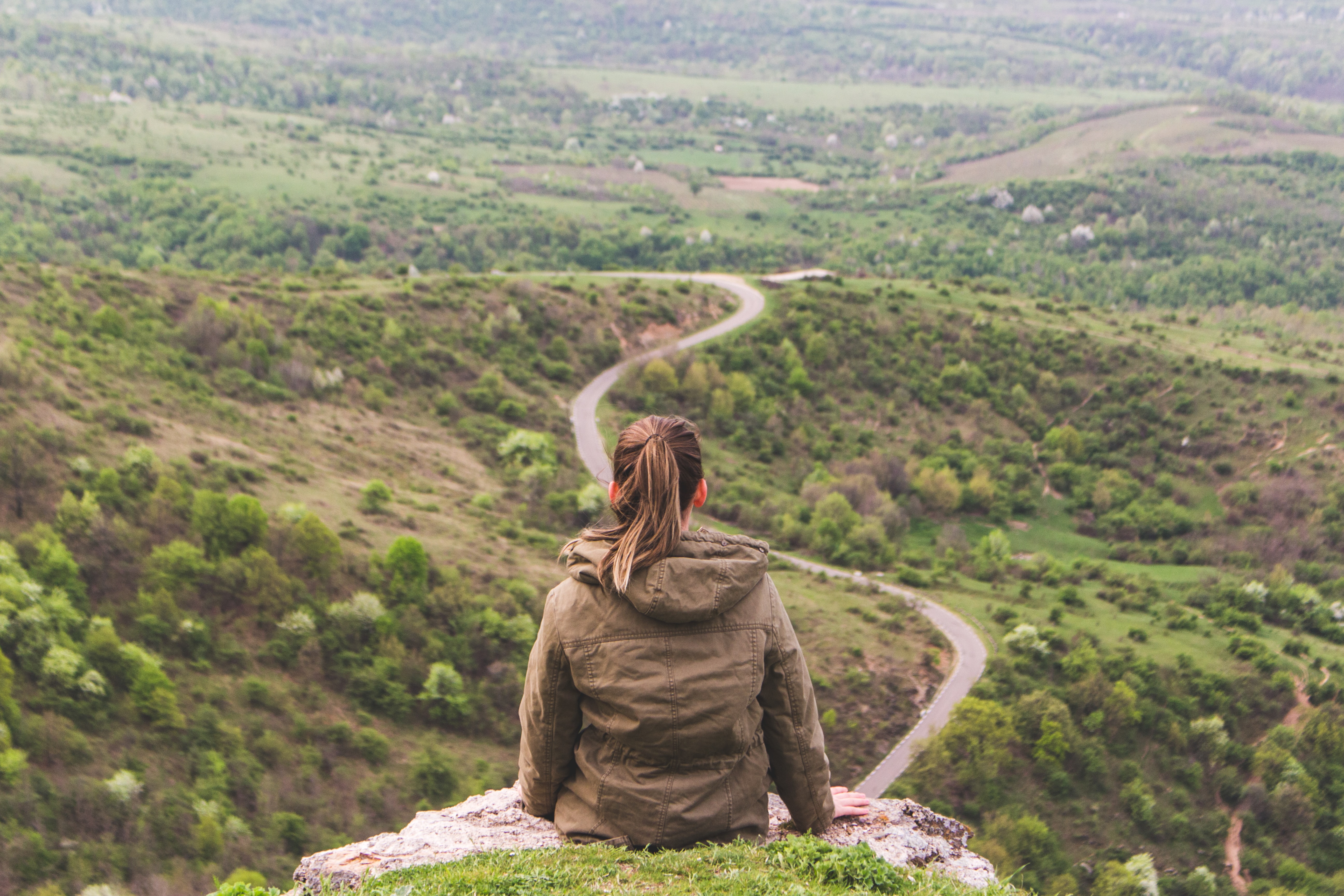 A young women sits on a cliff looking at a long winding road