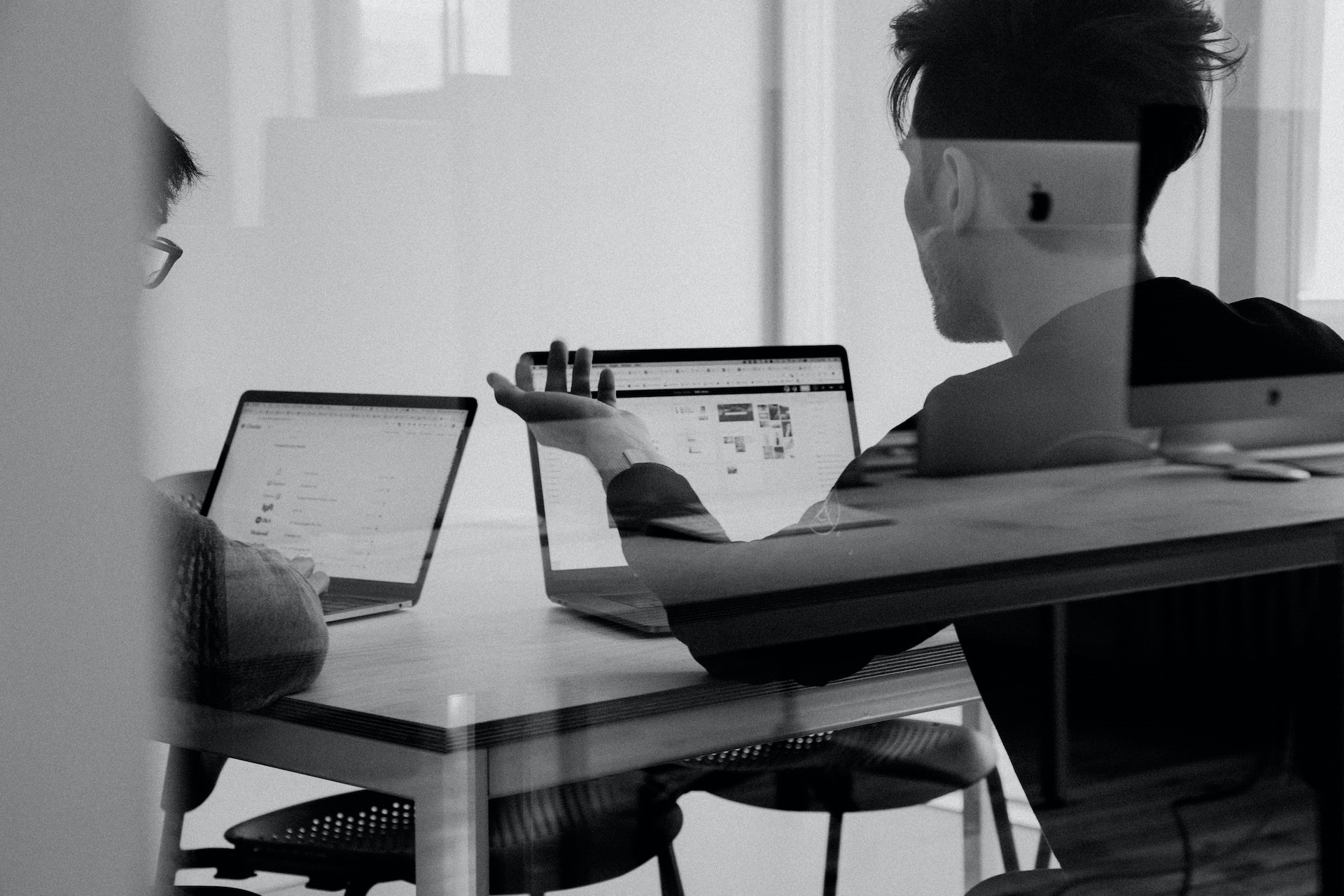 Black-and-white photo of two people, seen from behind, working on Apple laptops.