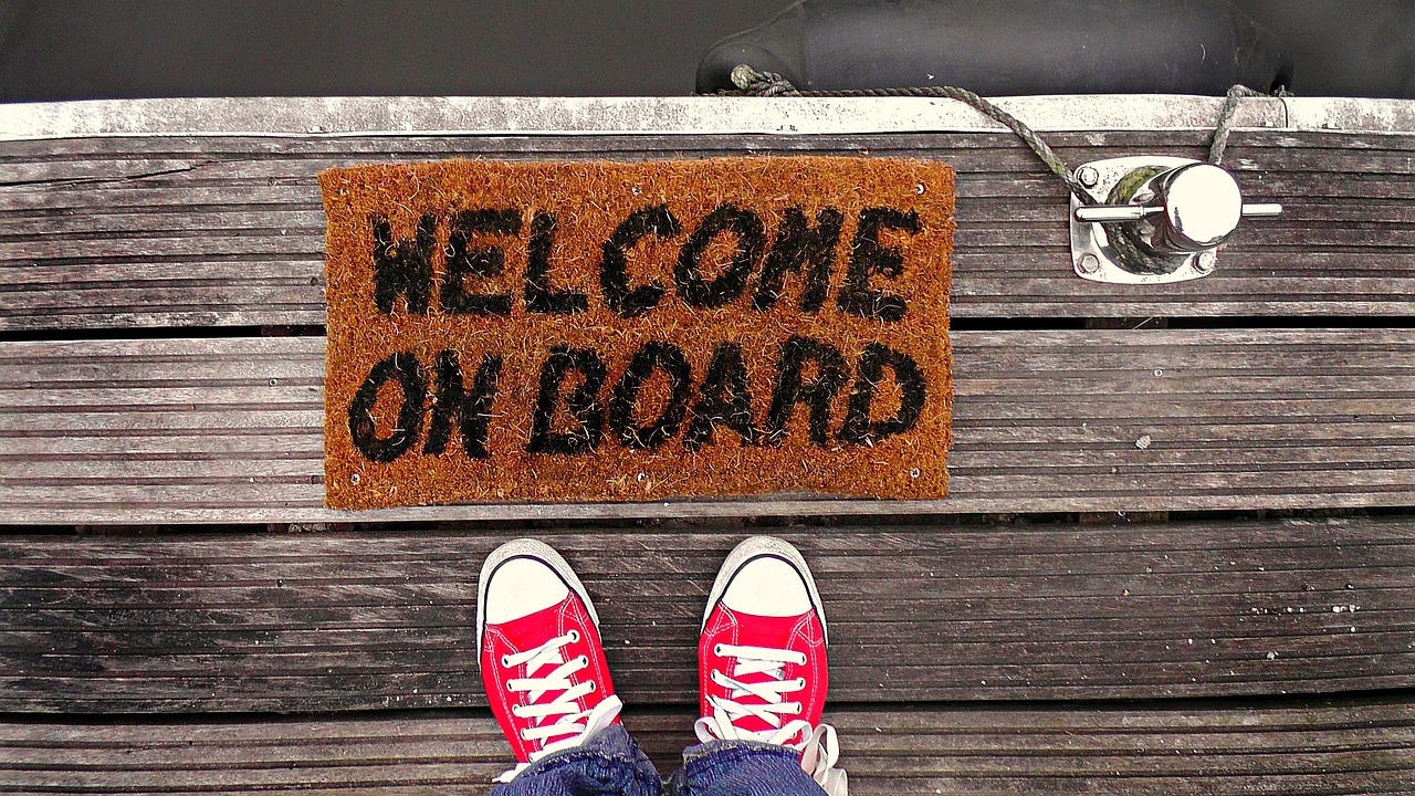 Image of a welcome mat on a jetty which reads 'Welcome on board'. So the bottom of the image we can see two red converse shoes.