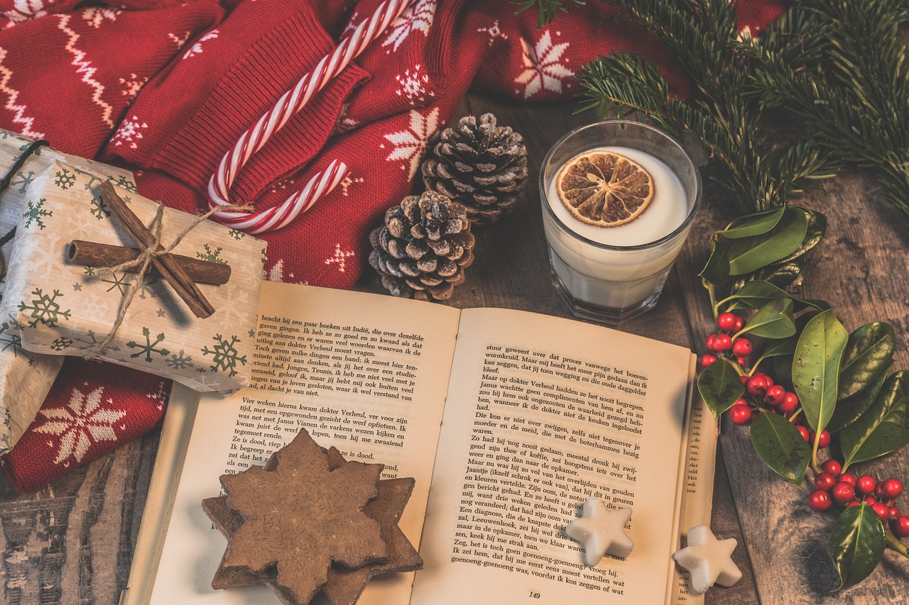 An open book surrounded by a collection of festive items, including eggnog, pine cones, candy canes and holly. The warm lighting and scattered items give a cozy feel. You can almost hear the crackling fire and smell the cinnamon.