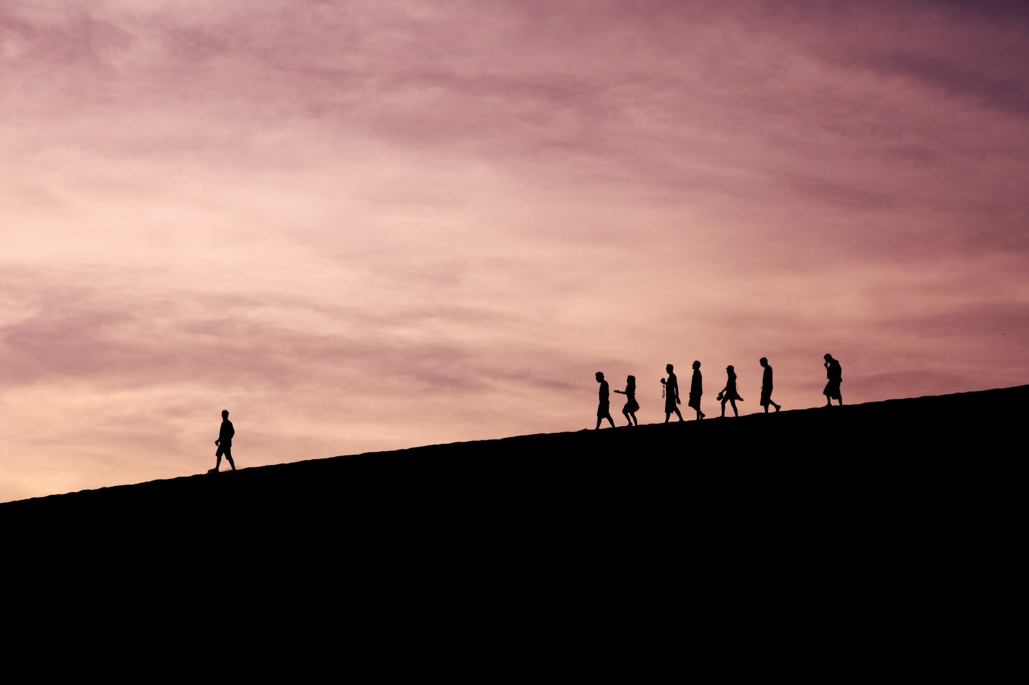 Silhouttes of a figure walking in front of a group of 7 people a few metres behind. The figures are walking down a hill set against a moody purple and orange, cloudy sunset.