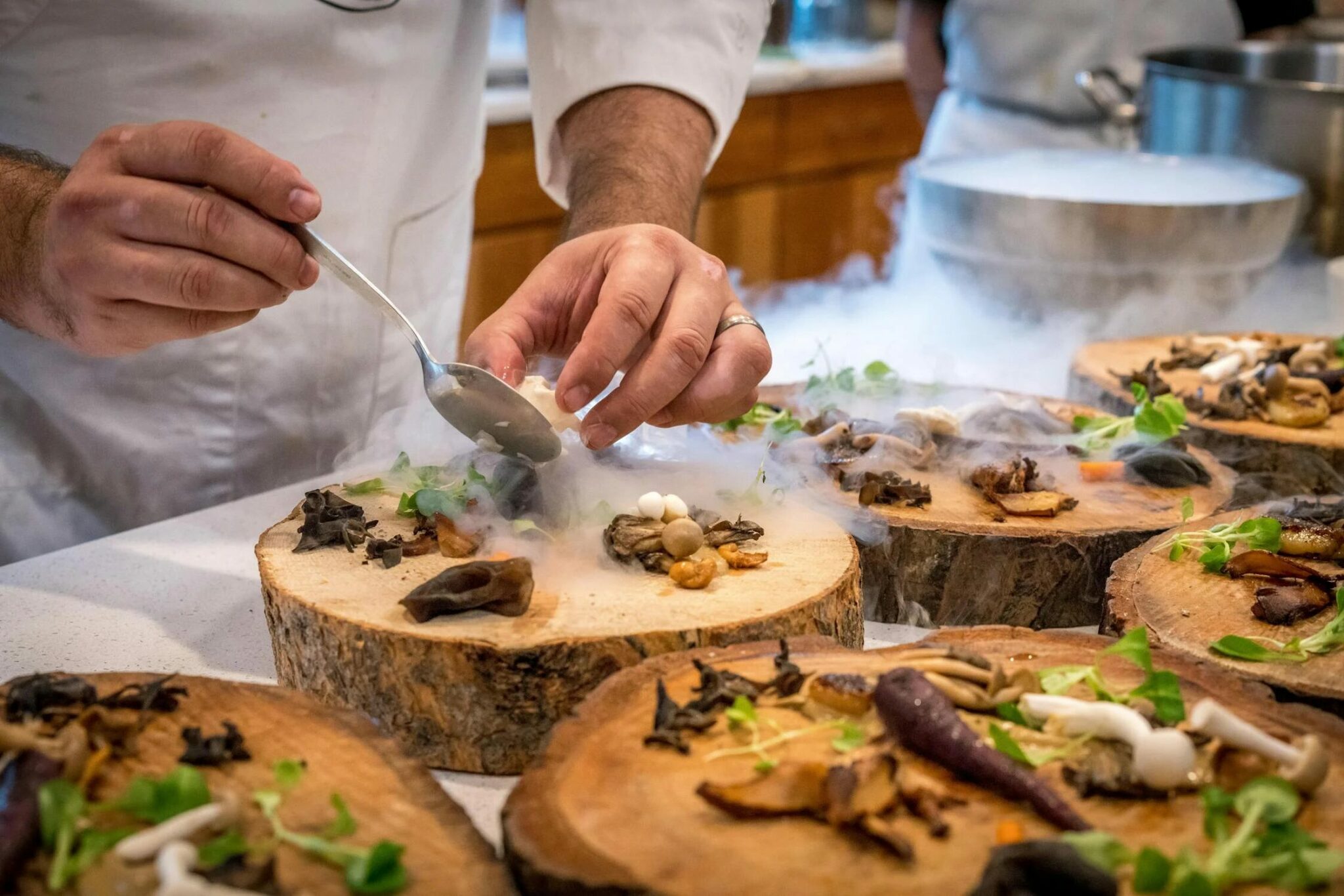 A photo of a chef's hands putting the finishing touches to a dish which is largely made up of mushrooms, and sits on a decorative piece of tree trunk.